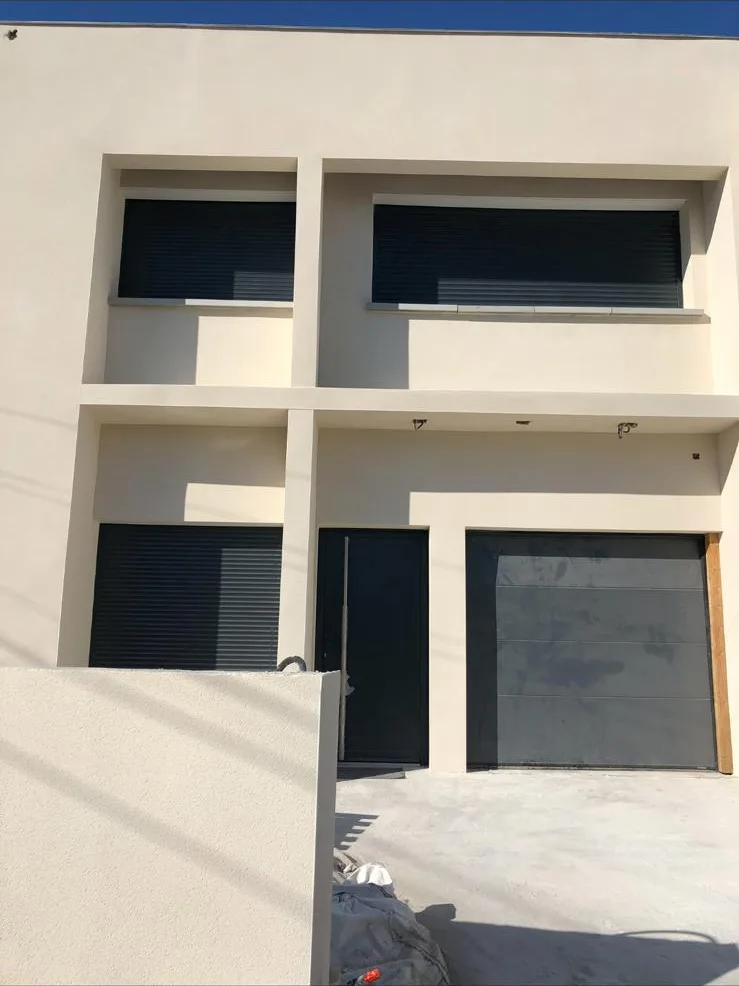 Maison neuve à l'enduit beige clair terminé, volets roulants anthracite et dalle béton au premier plan